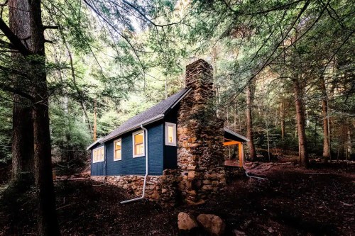 A cozy blue cabin nestled among tall trees, featuring a stone chimney and warm glowing windows.
