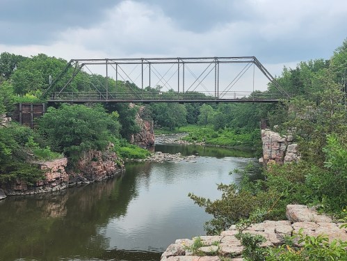 A rusted metal bridge spans a calm river, surrounded by lush greenery and rocky cliffs under a cloudy sky.