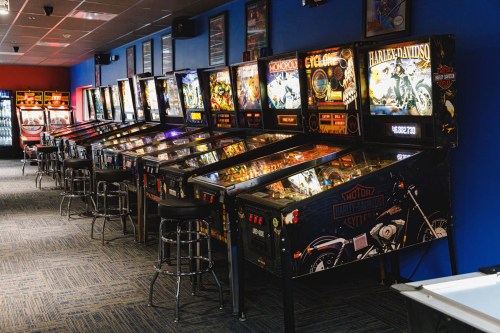 A row of vintage pinball machines in a brightly lit arcade with blue walls and bar stools.
