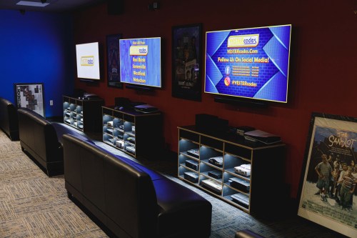 A cozy media room with black seating, multiple TVs displaying promotional content, and movie posters on the walls.