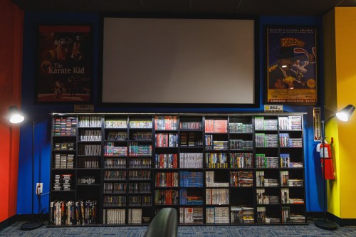 A cozy room with a large screen, colorful walls, and shelves filled with various video games and DVDs.