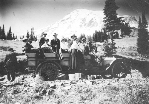 A vintage photo of a group of people in period clothing gathered around an old truck in a mountainous landscape.