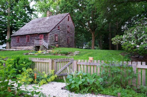 A rustic wooden house with red accents, surrounded by lush greenery and a garden, set in a peaceful outdoor setting.