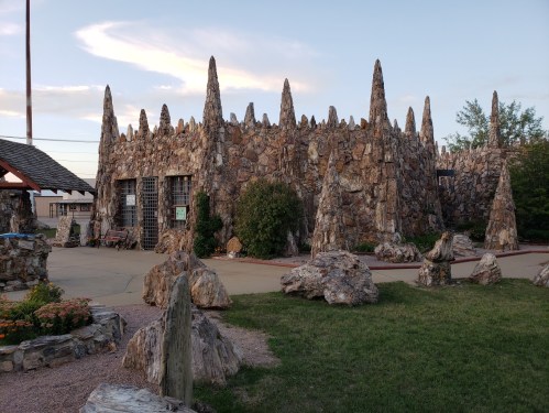 A unique stone building with spiky rock formations, surrounded by greenery and a paved area.