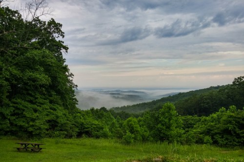 A misty valley view surrounded by lush green trees under a cloudy sky. A picnic table is visible in the foreground.