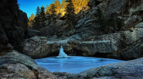 A serene rocky landscape with a frozen pond surrounded by trees and cliffs under a clear blue sky.