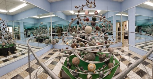A spiral sculpture with various colored spheres, surrounded by mirrors in a room with tiled flooring.