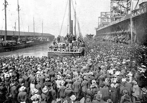 A large crowd gathers at a dock to welcome a boat, with ships and warehouses in the background.