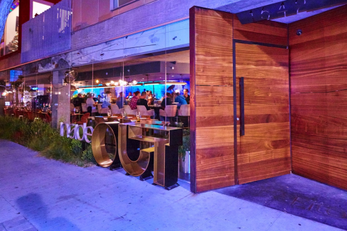 Exterior view of a modern restaurant with a wooden entrance and large glass windows, showcasing diners inside.