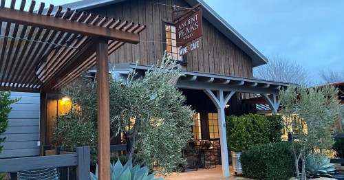 A rustic wooden building with a sign reading "Ancient Peaks Wine Cafe," surrounded by greenery and evening light.