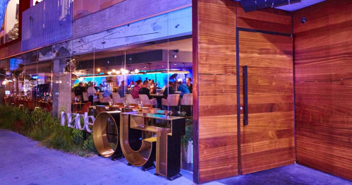 A modern restaurant entrance with wooden doors and a large "OUT" sign, featuring a lively dining area visible through glass.