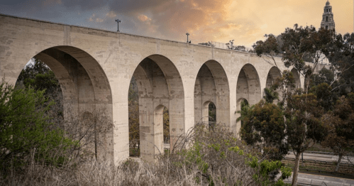 A stone aqueduct with multiple arches, surrounded by trees, under a dramatic sunset sky.