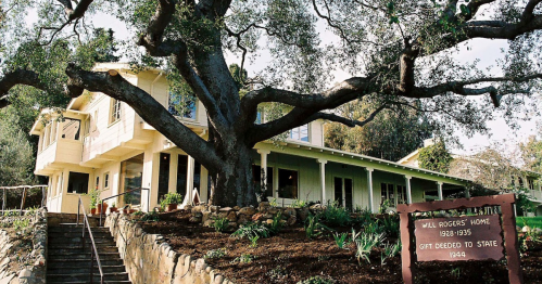 Historic Will Rogers Home, featuring a large tree and a sign, surrounded by greenery and steps leading to the entrance.