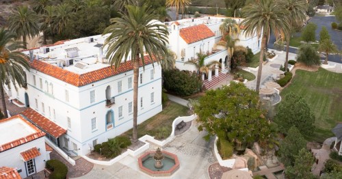 Aerial view of a white building with red tile roofs, surrounded by palm trees and landscaped gardens.