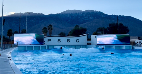 A wave pool with surfers riding waves, surrounded by mountains and large screens displaying surf visuals.