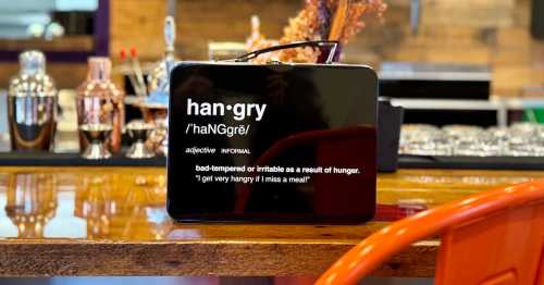 A black lunchbox with the word "hangry" and its definition displayed on the front, set on a bar counter.