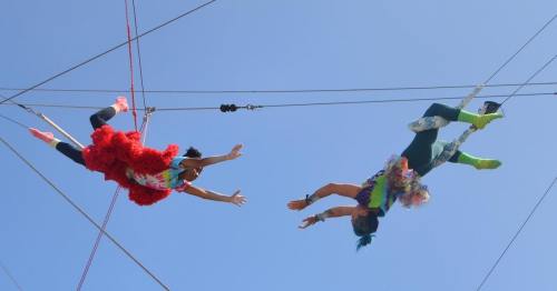 Two acrobats in colorful costumes perform a trapeze act against a clear blue sky.
