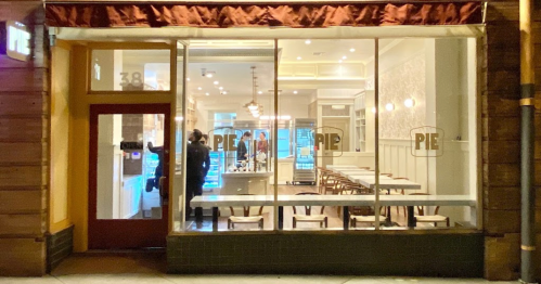 Exterior view of a restaurant named "PIE," featuring large windows and a cozy interior with tables and chairs.