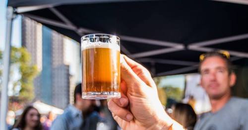 A hand holds up a glass of beer in a lively outdoor setting, with people and tents blurred in the background.