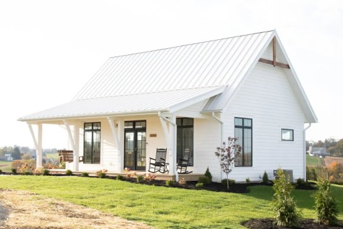 A modern white house with a metal roof, large windows, and a porch featuring rocking chairs, surrounded by greenery.