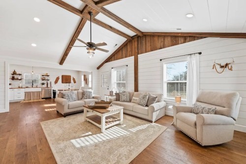 Bright, modern living room with white walls, wooden beams, cozy furniture, and a large area rug. Natural light floods the space.