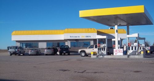 A gas station with a yellow roof, several cars parked, and a sign indicating diesel fuel availability.