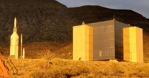 A rocket stands beside a modern glass building against a mountainous backdrop, illuminated by warm sunlight.