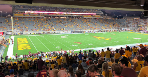 A view of a nearly empty football stadium with fans in yellow and maroon, under evening lights.