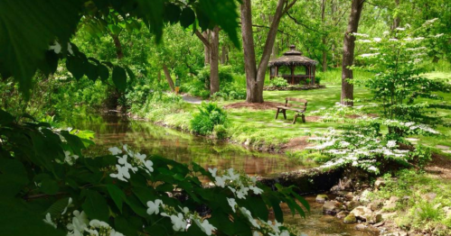 A serene garden scene with a gazebo, lush greenery, and a gentle stream, framed by blooming flowers.