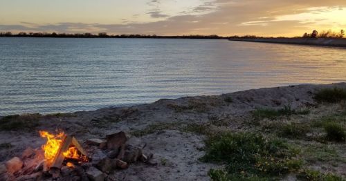 A serene lakeside scene at sunset, featuring a small campfire on the sandy shore with calm water in the background.