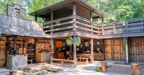 A rustic outdoor kitchen with wooden structures, a dining area, and a stone fire pit surrounded by greenery.