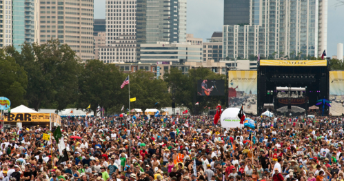 A large crowd at a music festival with city skyscrapers in the background and a stage visible.