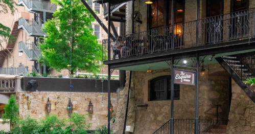 A rustic building with a balcony, surrounded by greenery and decorative lights, featuring outdoor seating.