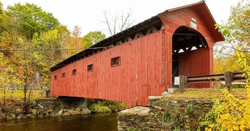 A red covered bridge spans a calm river, surrounded by autumn foliage and trees.