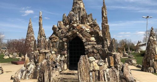 A rocky, castle-like structure with tall spires and an archway, surrounded by a paved area and blue sky.