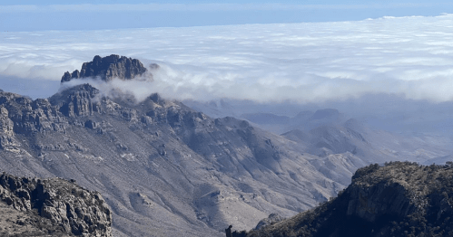 A panoramic view of rugged mountains with clouds rolling over the peaks, creating a dramatic landscape.