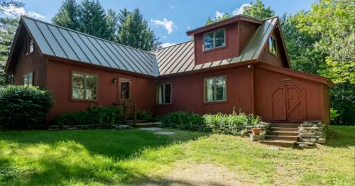 A red house with a metal roof, surrounded by greenery and trees, featuring a front porch and stone steps.
