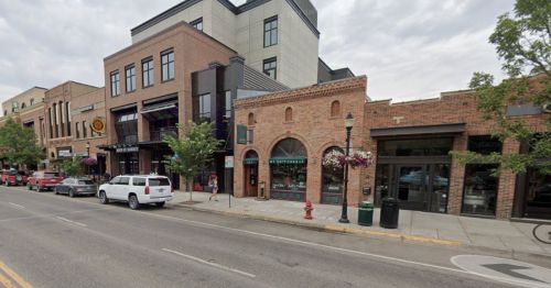 A street view of a downtown area featuring brick buildings, shops, and parked cars under a cloudy sky.
