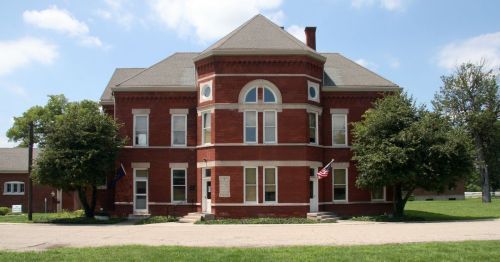 Historic red brick building with large windows, surrounded by trees and a grassy area, under a blue sky.