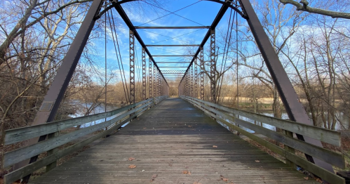 A metal bridge spans a river, surrounded by bare trees and a clear blue sky. The wooden walkway leads into the distance.