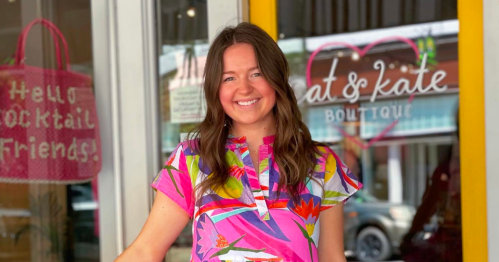 A smiling woman in a colorful floral dress stands in front of a boutique with a cheerful sign.