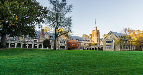 A historic stone building with a tower, surrounded by green grass and trees, under a clear blue sky.
