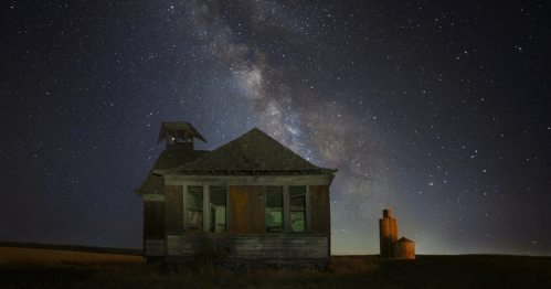 A dilapidated house under a starry sky, with the Milky Way visible and a silhouette of a nearby structure.
