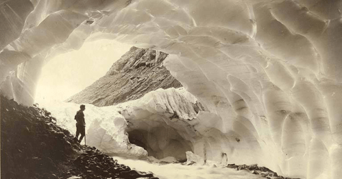 A person stands inside a glacial cave, surrounded by ice formations and rocky terrain.
