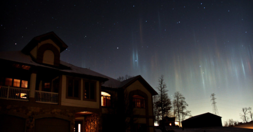A house under a starry night sky with colorful auroras and silhouettes of trees in the foreground.