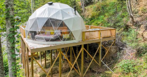 A geodesic dome house on stilts, surrounded by trees, with a wooden deck and large windows.