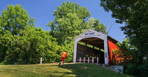 A person walks towards a historic covered bridge surrounded by lush green trees on a sunny day.
