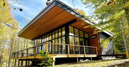Modern cabin with large windows and a wooden deck, surrounded by trees and autumn foliage.