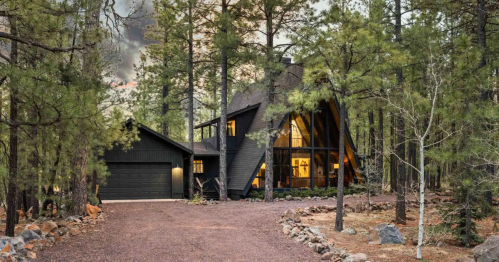 A modern A-frame house surrounded by tall pine trees, with a gravel driveway and warm lights glowing from the windows.