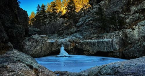 A frozen pond surrounded by rocky cliffs and evergreen trees, illuminated by warm sunlight in the background.